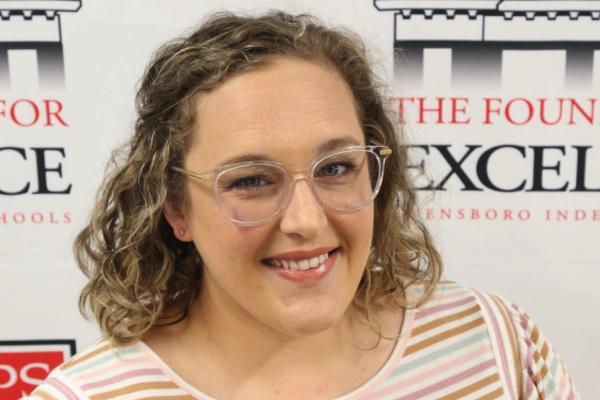 Photo from the shoulders up of a woman with shoulder length hair and glasses in front of a backdrop with text that says "The foundation for excellence."